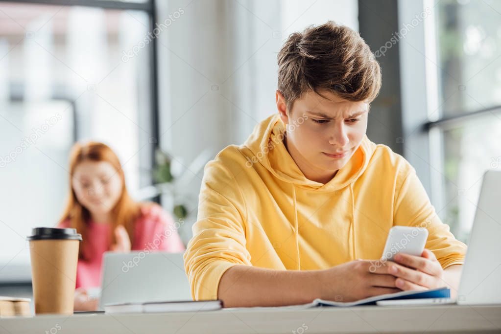 pensive boy in yellow hoodie using smartphone at desk