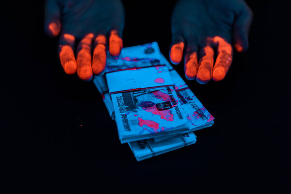 cropped view of man with orange fingerprints near russian money under uv lighting isolated on black 