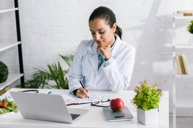 attractive woman in white coat writing in notebook near laptop and vegetables  clipart