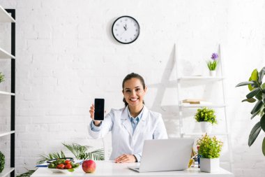 happy nutritionist holding smartphone with blank screen near laptop  clipart
