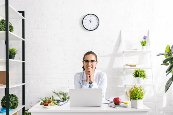cheerful nutritionist in white coat near vegetables and laptop 