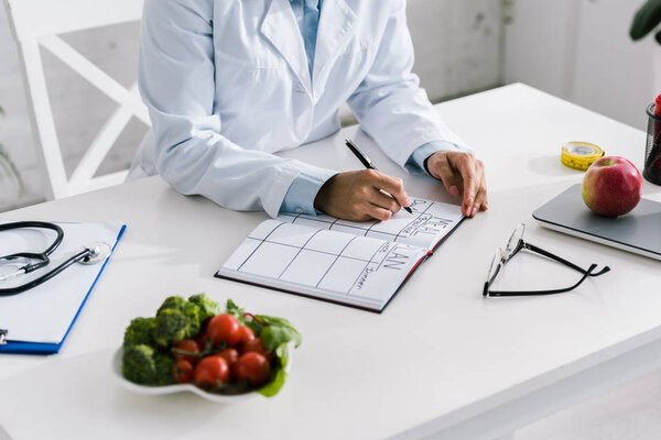 cropped view of nutritionist writing in notebook near vegetables and apple 