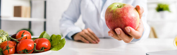 panoramic shot of nutritionist holding tasty apple near vegetables 