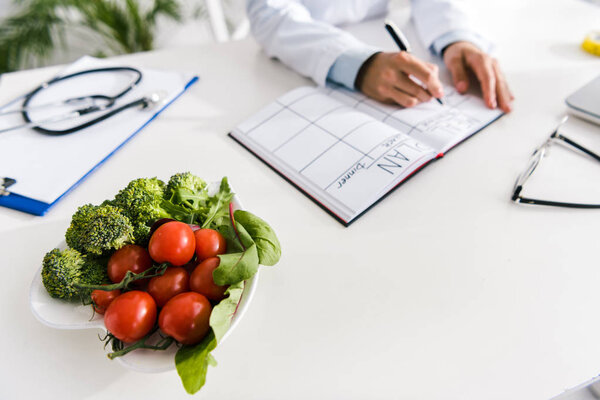 cropped view of nutritionist writing in notebook with meal plan near vegetables 