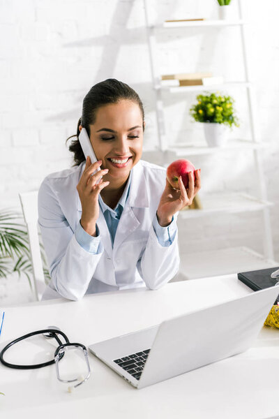 cheerful nutritionist talking on smartphone and holding apple in clinic 