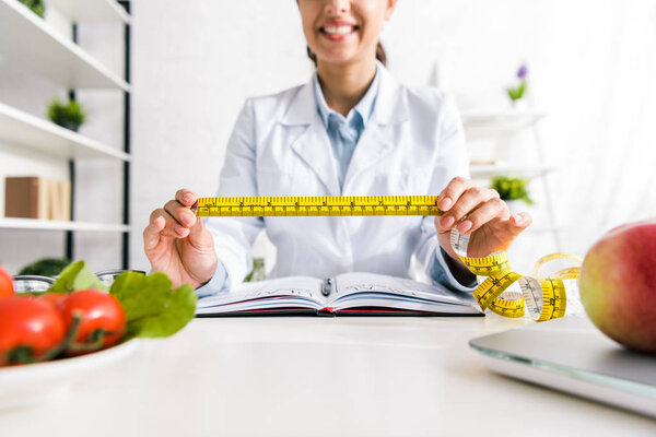 cropped view of cheerful woman holding measuring tape near vegetables and apple 