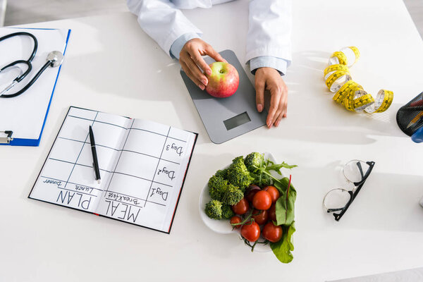 top view of nutritionist holding apple on food scales near vegetables 