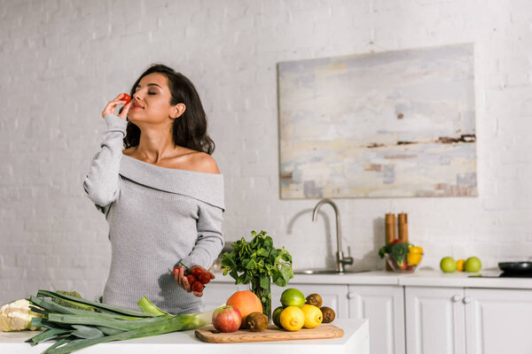 attractive girl smelling cherry tomato near vegetables 