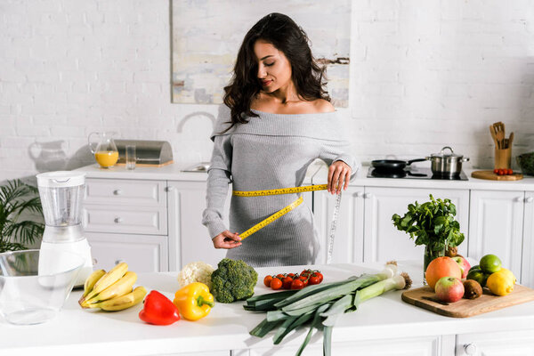 attractive woman measuring waist near fruits and vegetables on table  