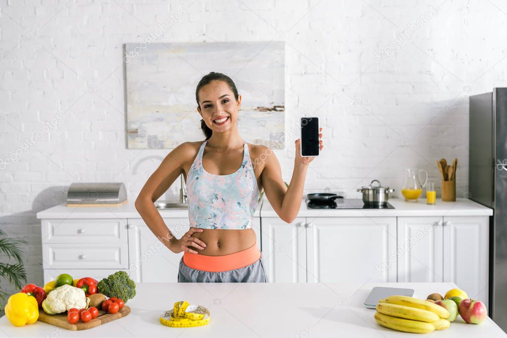 cheerful woman holding smartphone with blank screen and standing with hand on hip 