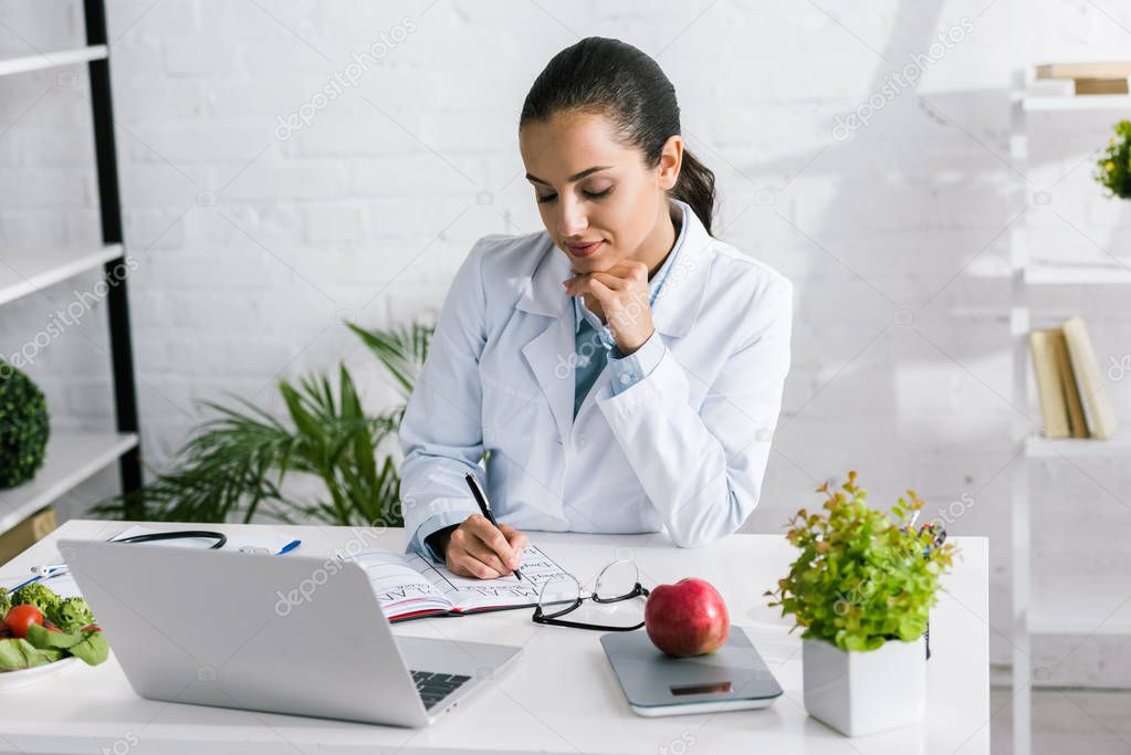 attractive woman in white coat writing in notebook near laptop and vegetables 