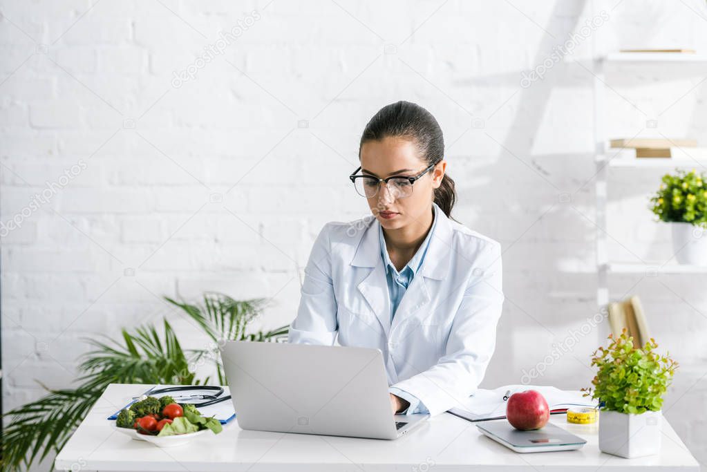 attractive nutritionist in glasses using laptop near vegetables and plants 