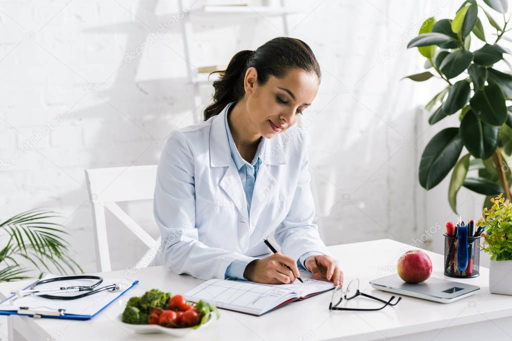 attractive young nutritionist in white coat writing in notebook 