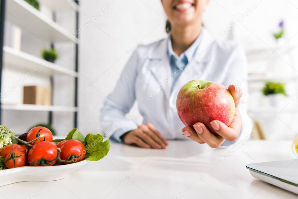 cropped view of nutritionist holding tasty apple near vegetables 
