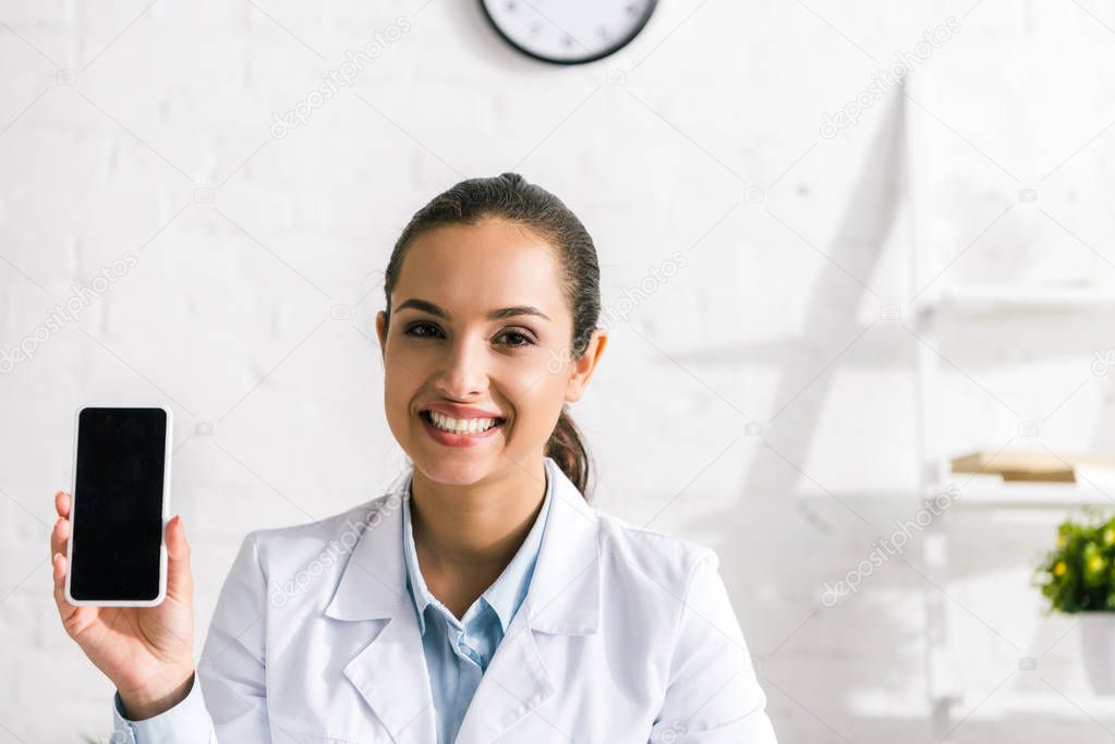 happy doctor in white coat looking at camera and holding smartphone with blank screen 
