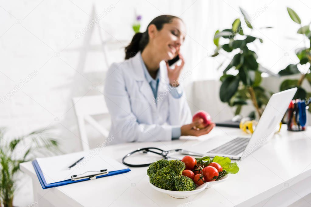 selective focus of vegetables near happy nutritionist talking on smartphone 