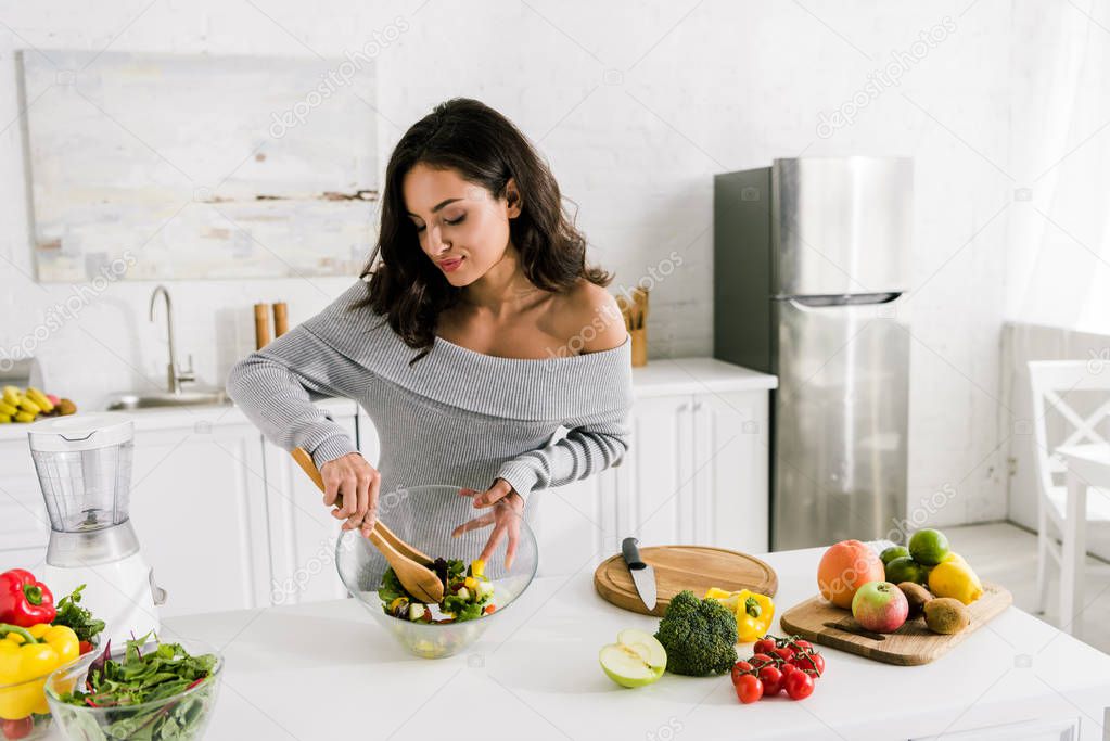 happy woman mixing fresh salad near vegetables at home 