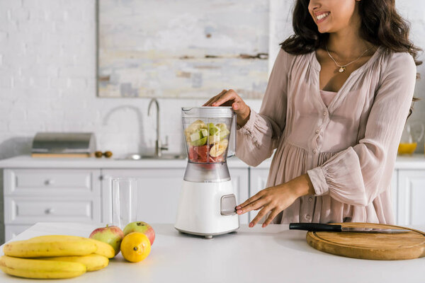 cropped view of happy pregnant woman touching button on blender with fruits 