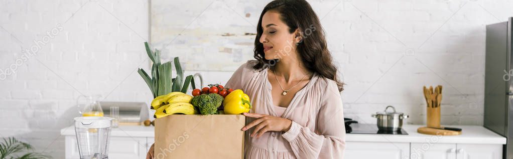 panoramic shot of pregnant woman looking at paper bag with groceries 