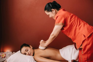 side view of asian masseur doing back massage with herbal balls to woman in spa clipart