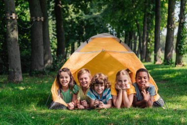 happy multicultural kids smiling while lying near camp  clipart