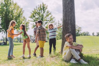 ağaç gövdesi ve çok kültürlü çocukların yanında oturan üzgün çocuğun seçici odak 