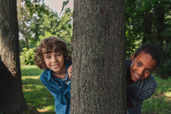 木の幹とアフリカ系アメリカ人の友人の近くにカメラを見てかわいいカーリー少年 — ストック写真