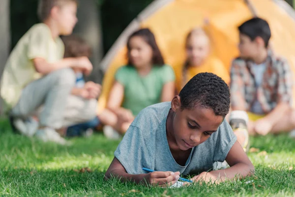 Foco Selectivo Niño Afroamericano Feliz Escribiendo Cuaderno Cerca Amigos Campamento — Foto de Stock