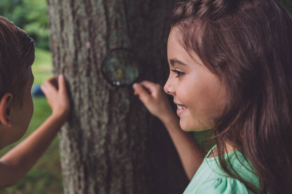 selective focus of happy kid holding magnifier while standing near tree trunk with friend 