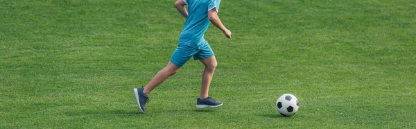 panoramic shit of kid in sportswear running on green grass with football 