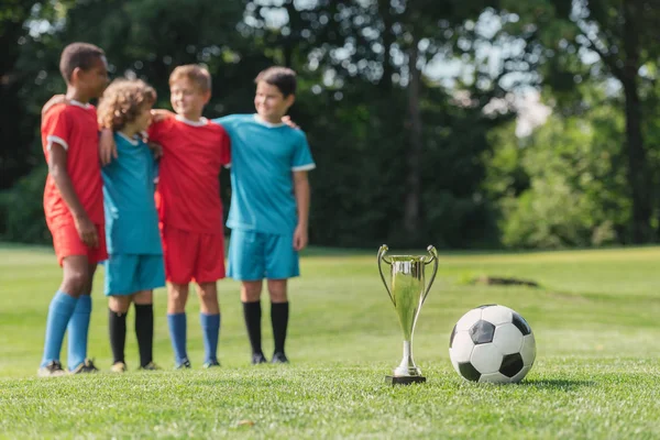 selective focus of trophy and football near cute multicultural winners hugging outside 