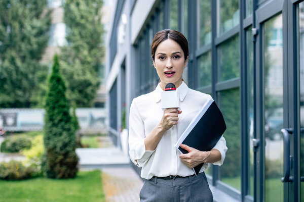 attractive journalist in formal wear holding microphone and speaking outside 