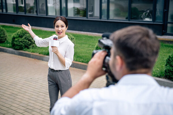 selective focus of attractive journalist holding microphone and cameraman shooting her outside 