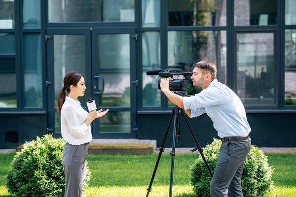 side view of attractive journalist holding microphone and cameraman shooting her outside 