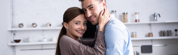 panoramic shot of smiling couple embracing and looking at camera at home