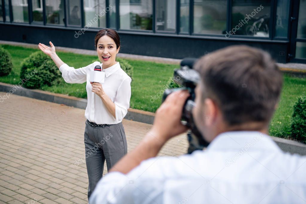 selective focus of attractive journalist holding microphone and cameraman shooting her outside 
