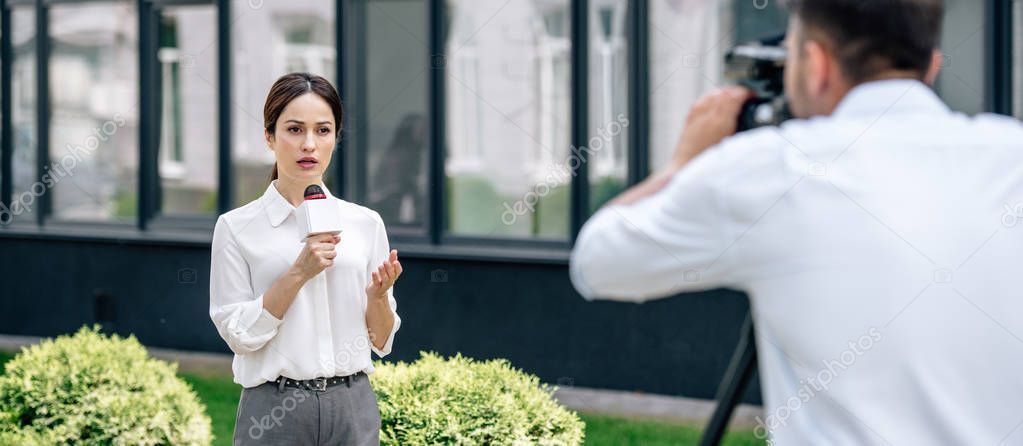 panoramic shot of attractive journalist holding microphone and cameraman shooting her outside 