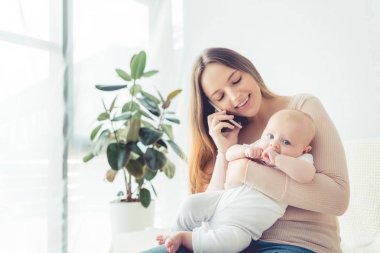 çekici ve gülümseyen anne çocuğunu tutarak ve dairede akıllı telefon konuşurken 