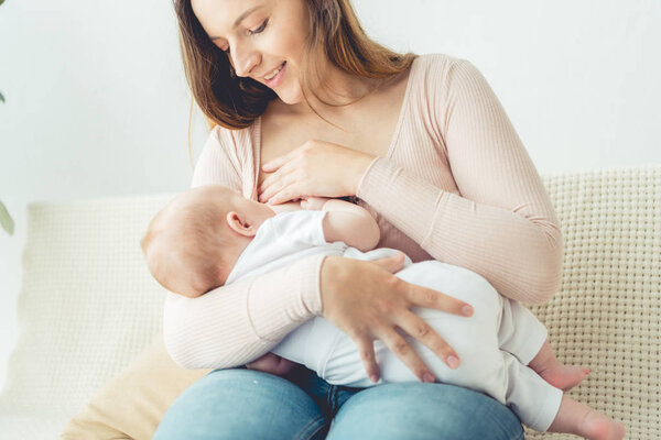 attractive and smiling mother breastfeeding her child in apartment 