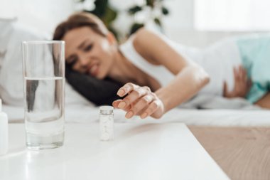 selective focus of suffering woman in bed and pills with glass of water on foreground clipart