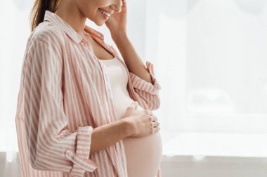 partial view of happy pregnant woman smiling and touching belly clipart