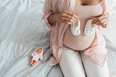 yatakta oturan ve bebek ayakkabıları tutan hamile kadın kırpılmış görünümü