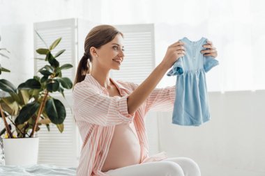 mutlu gülümseyen hamile kadın yatakta oturan ve bebek giysileri tutan