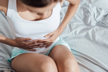 yatakta oturan ve karnına dokunan genç kadının kırpılmış görünümü