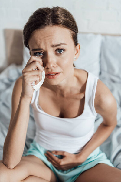 high angle view of stressed young woman touching belly and crying in bed
