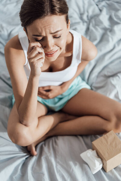 high angle view of stressed young woman touching belly and crying in bed
