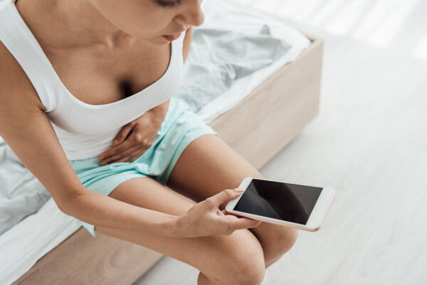 cropped view of young woman touching belly and using smartphone in bedroom