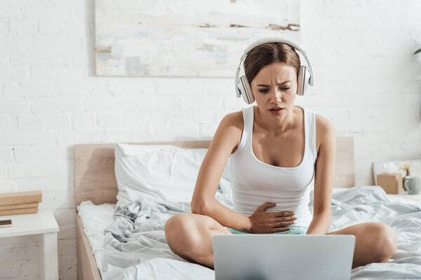 stressed woman in headphones touching belly and using laptop on bed