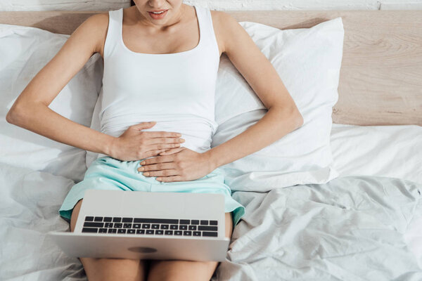 partial view of stressed woman touching belly while using laptop in bed