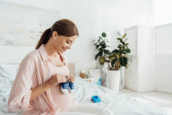 Uśmiechnięta Kobieta Ciąży Siedzi Łóżku Trzyma Buty Dla Niemowląt — Zdjęcie stockowe